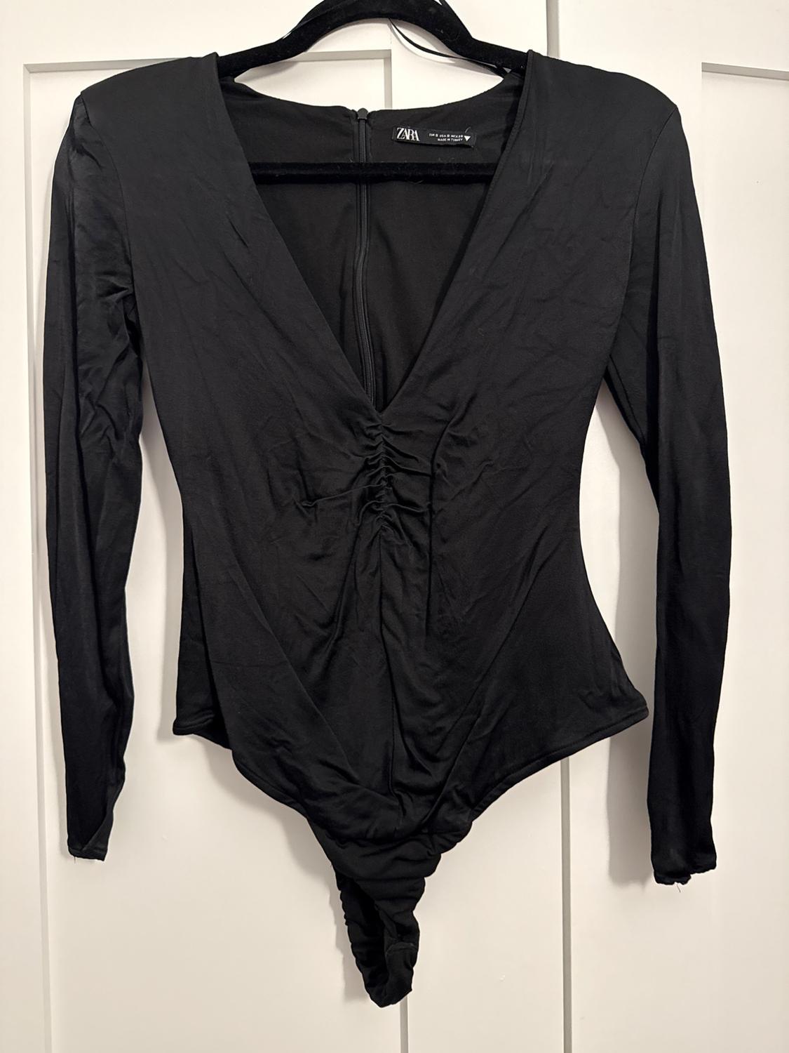 Black long-sleeved bodysuit with a V-neck and ruched front, hanging on a black hanger against a white background.