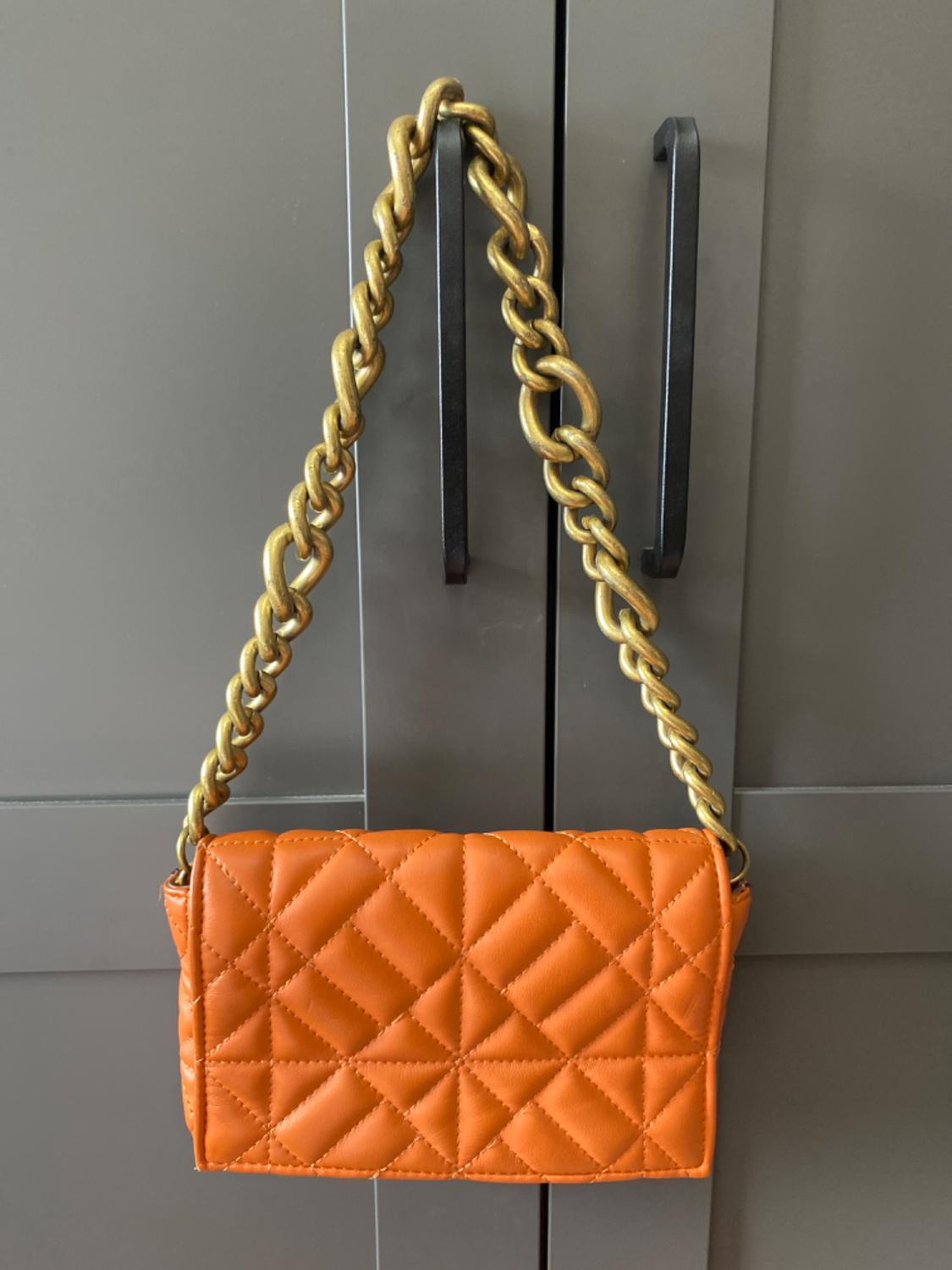 Orange quilted handbag with a gold chain, hanging from a grey wardrobe with black handles.