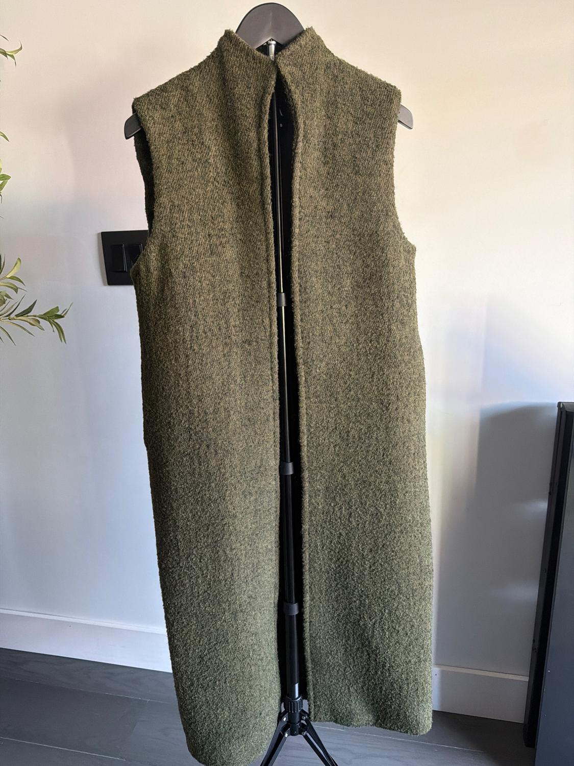 Olive green sleeveless long wool vest on a black hanger, white background with decorative plant on the left.