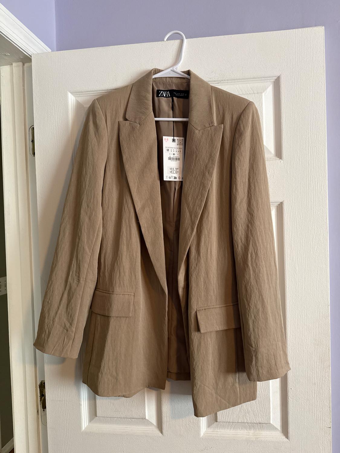 Beige jacket hanging on a white door, with a visible Zara tag. Indoor setting with a lilac wall in the background.