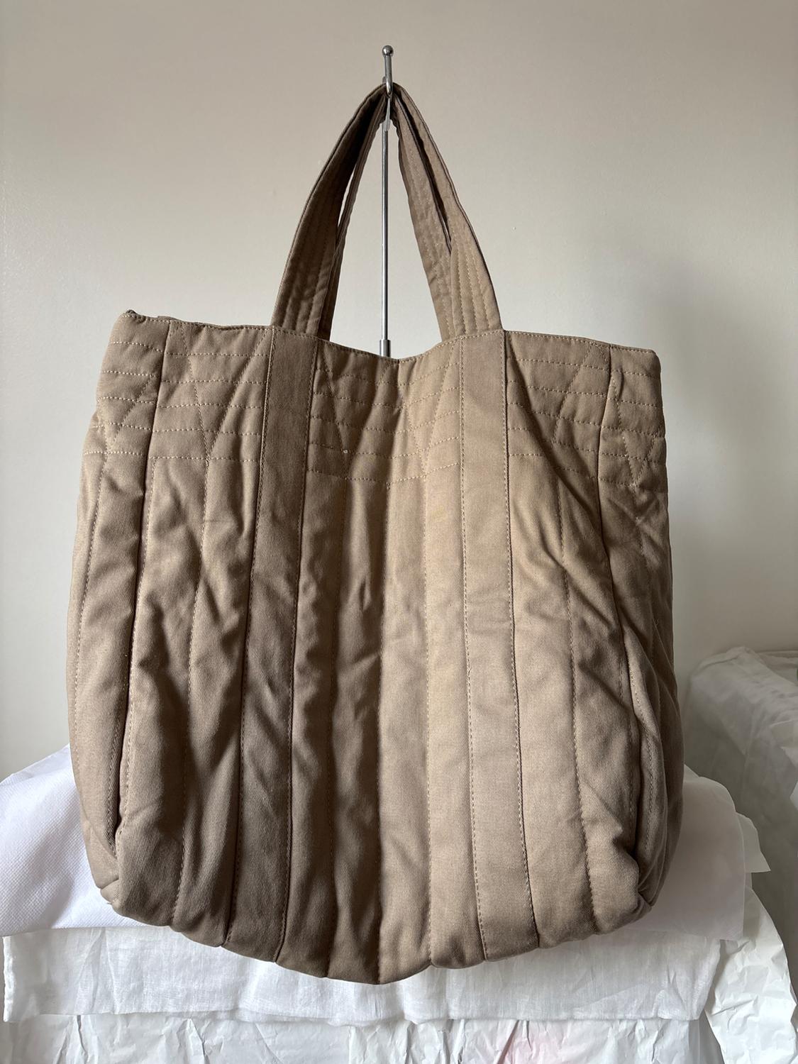 Large, light brown quilted fabric bag with short handles, on a neutral background and resting on a wrinkled white surface.