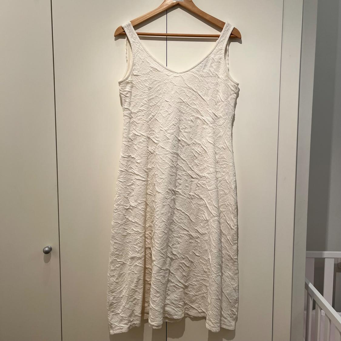 White sleeveless dress in a crinkle-texture fabric, hanging on a wooden hanger in front of a white wardrobe.