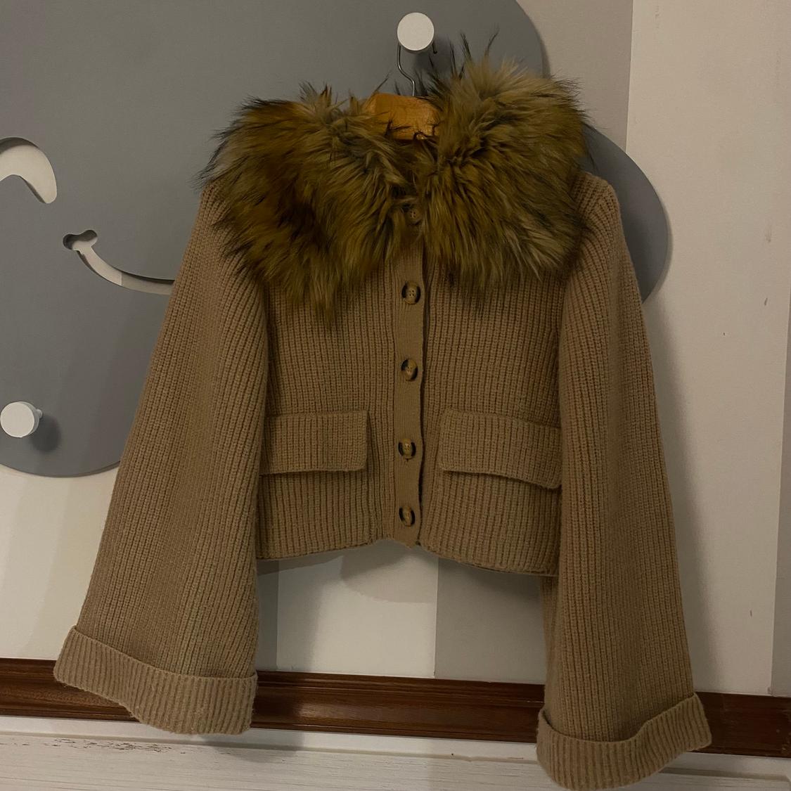 Beige knitted cardigan with buttons and a brown faux fur collar, hanging on a hanger against a grey and white background.