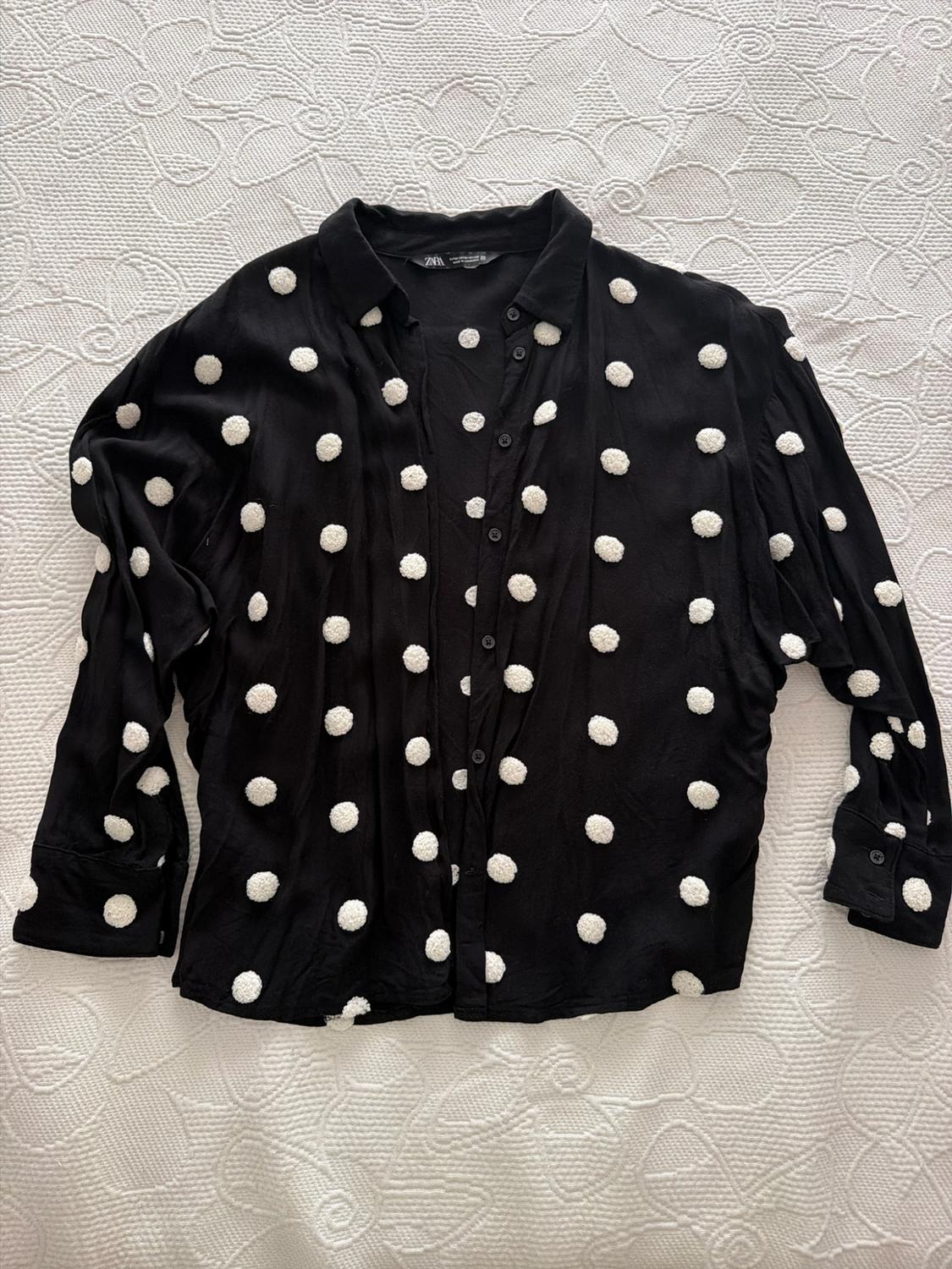 Black button-down shirt with raised white polka dots, on a white bedspread with a floral pattern. Relaxed style. Visible text: ZARA.