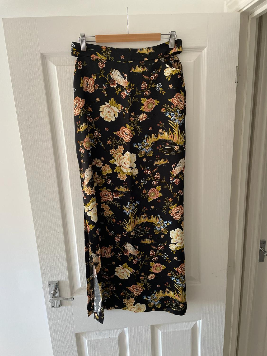 Long black skirt with a floral and bird print, featuring a side slit. Hanging on a hanger on a white door.