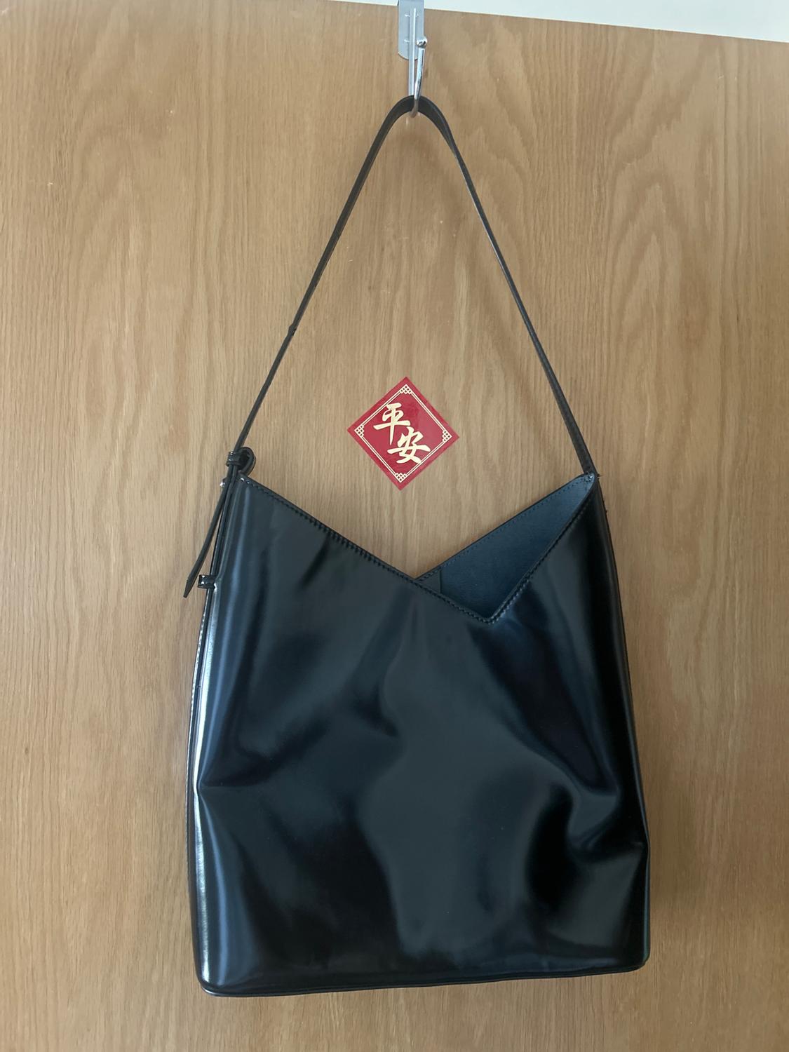 Shiny black shoulder bag with an asymmetric edge hanging on a wooden door. Behind it is a red ornament with the text 平安.