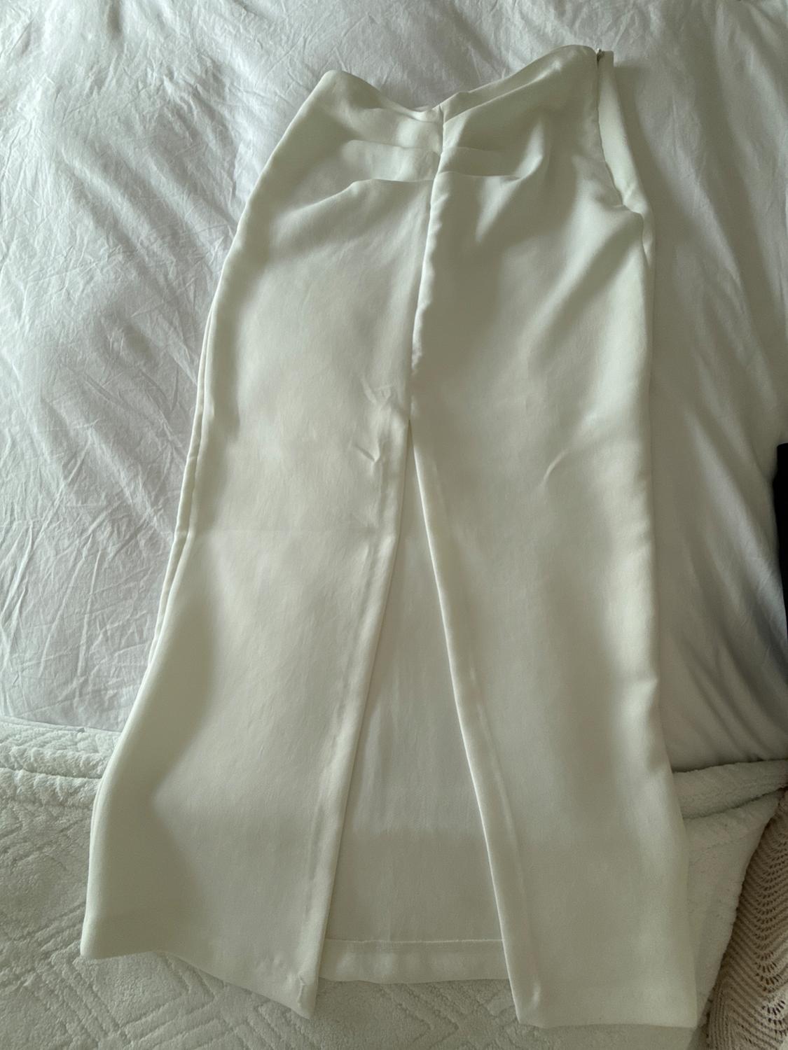 White lightweight fabric pants with pleats, placed on a bed with white sheets.