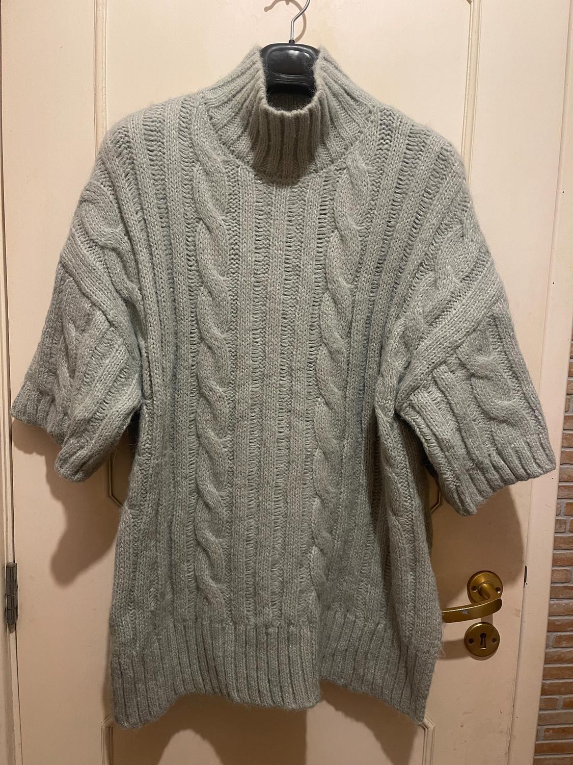 Chunky knit grey jumper with a high neck and cable-knit design, hanging on a hanger over a beige door.