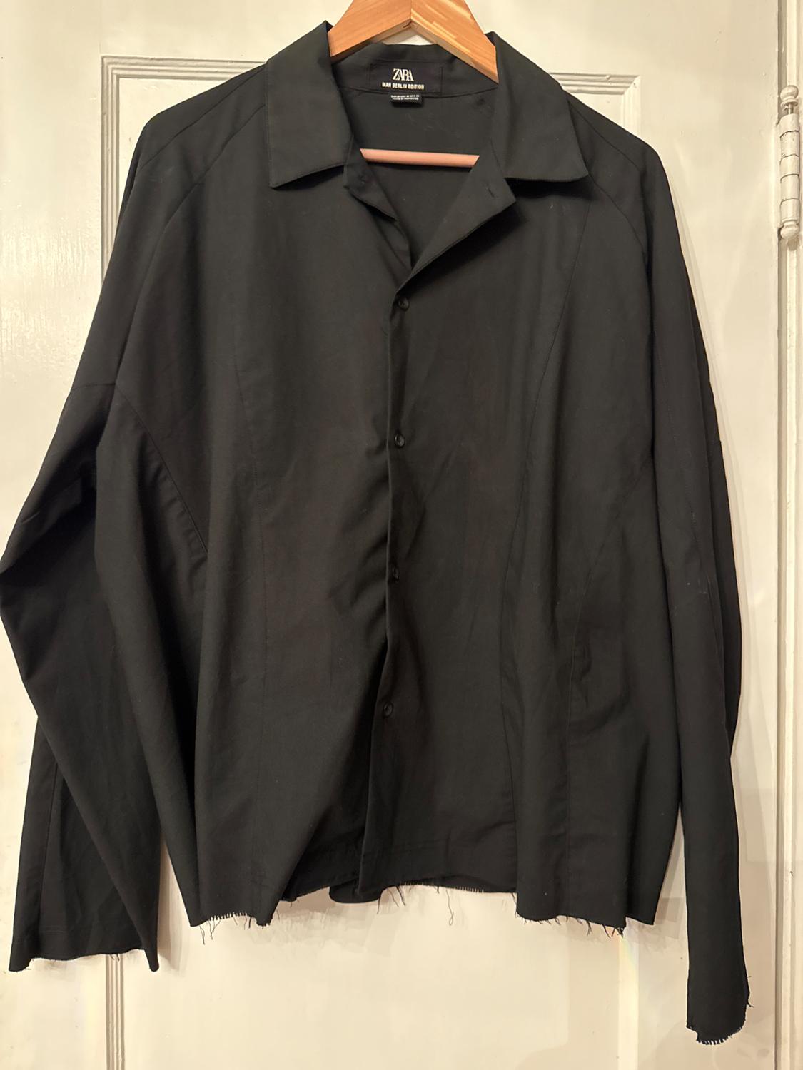 Black long-sleeved shirt hanging on a wooden hanger, with a frayed hem and a white door background.