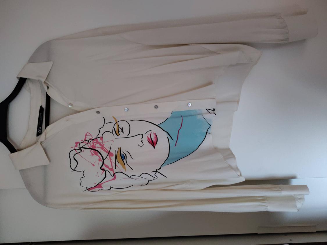 White hanging shirt with an abstract face design in blue, pink, and yellow. White background.