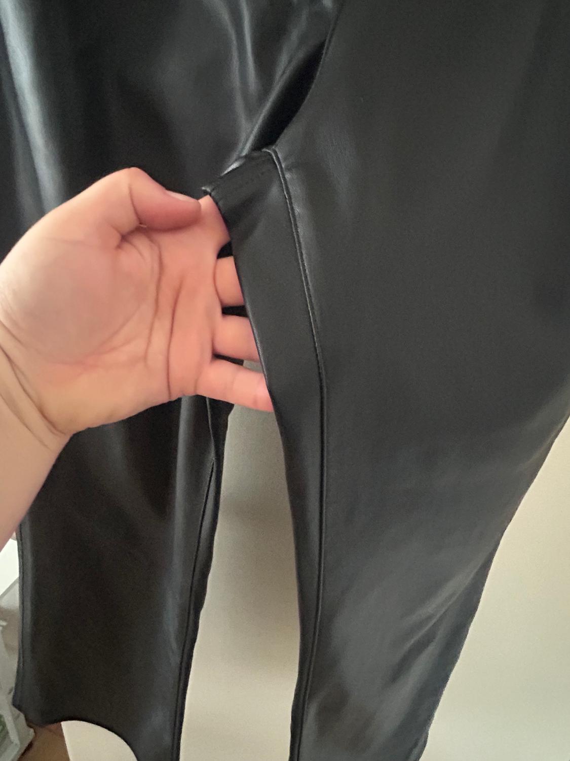Hand holding black trousers made of shiny material, showing seam detail and texture. Neutral background.