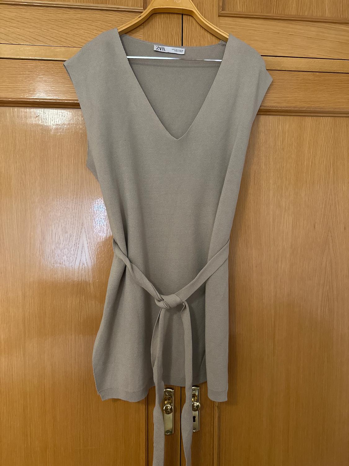 Beige sleeveless dress with a V-neck and belt, hanging on a wooden hanger over a wooden door.
