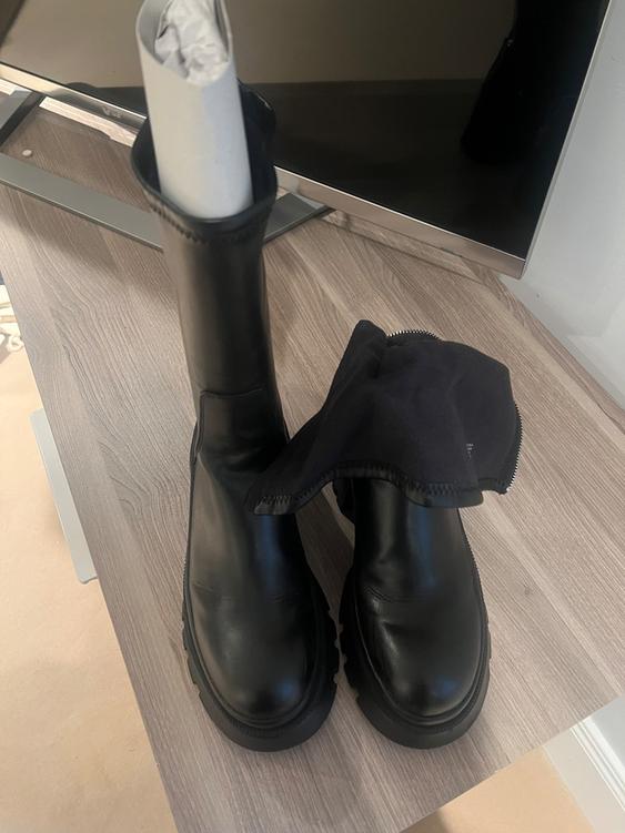 Black leather boots on a light wooden table, one with rolled paper inside and the other partially covered with black fabric.