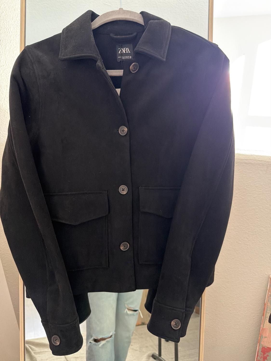 Black jacket hanging on a rail in front of a mirror. Reflection of ripped jeans and visible tag with "ZARA" text. Lit environment.