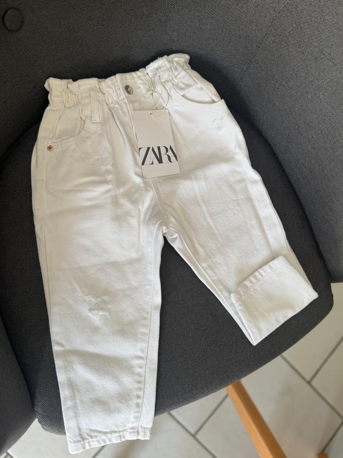 White trousers with an elasticated gathered waist, distressed details and a turn-up hem on a grey chair. Label with the text ZARA.