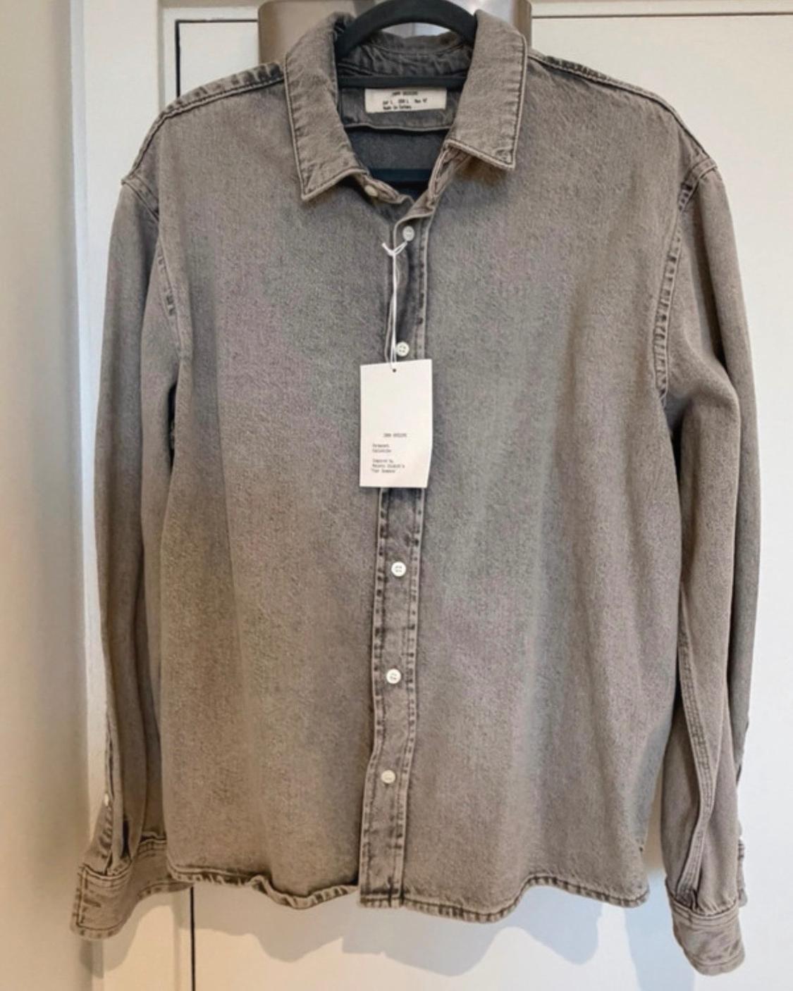 Grey long-sleeved shirt with a washed effect, buttoned up and hanging on a black hanger in front of a white door. It has a tag hanging from it.