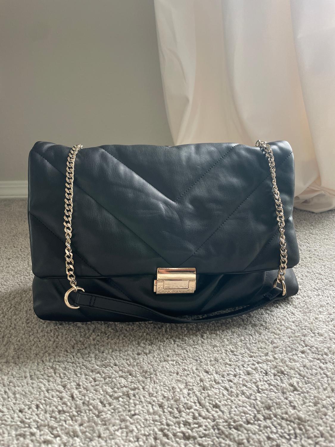 Black quilted handbag with gold chain on grey rug, white curtain in background.