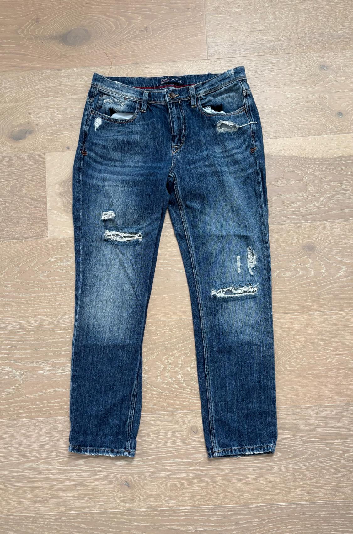 Blue denim jeans with a worn-in finish and rips, placed on a light wood floor. Z1975 DENIM interior label.