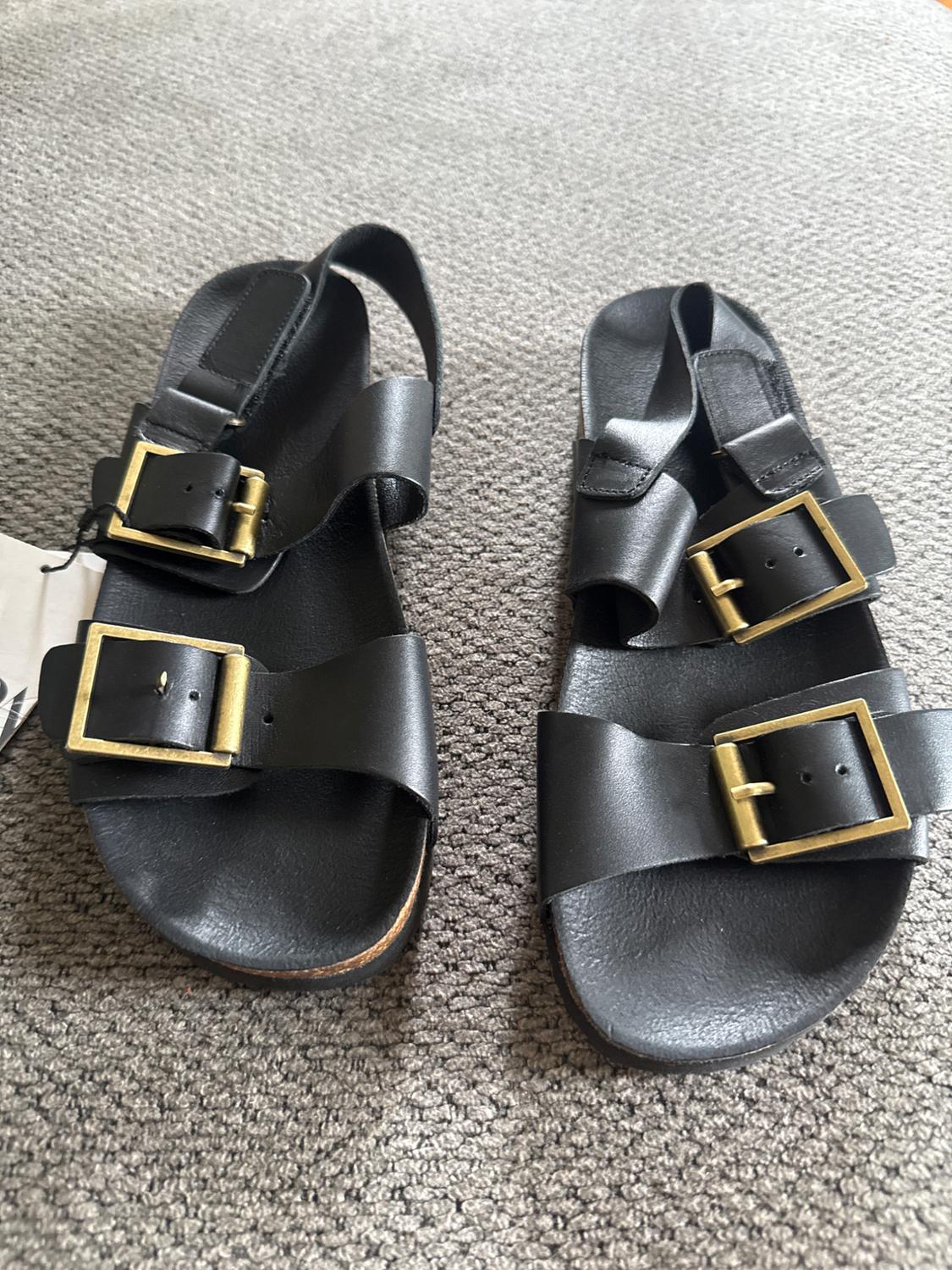 Black leather sandals with two adjustable straps and large gold buckles, placed on a grey rug with visible texture.