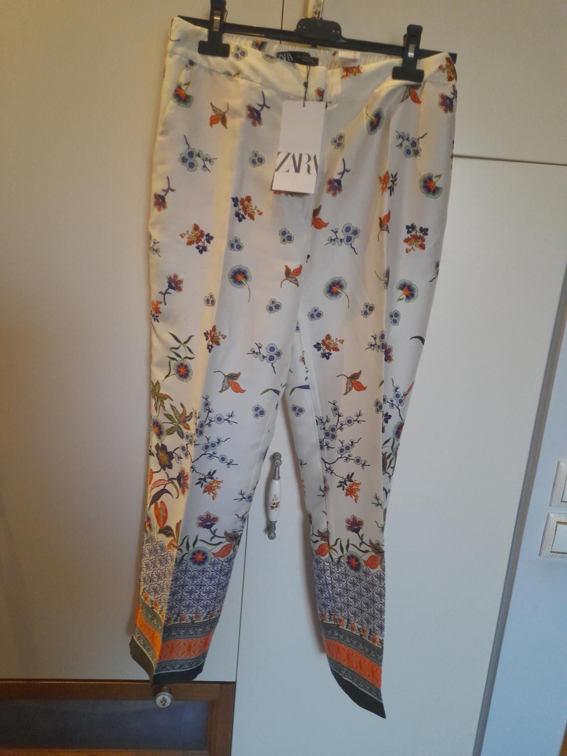Colourful floral print trousers with geometric borders. Visible label with the text "ZARA". White wardrobe background.