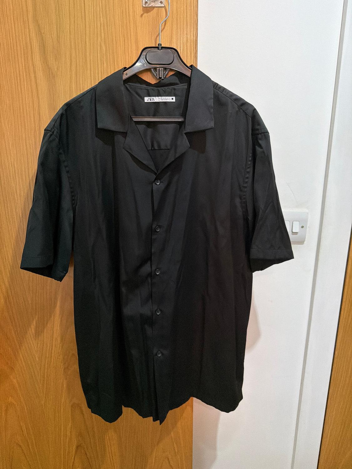Black short-sleeved shirt hanging on a hanger over a light wooden door. Label visible on the collar.