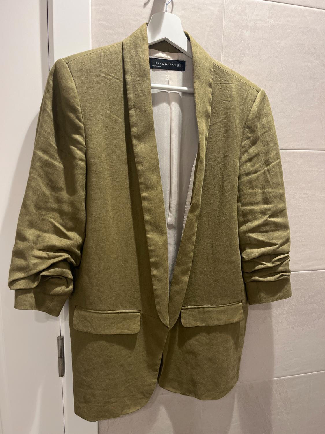 Olive green women's jacket hanging on a white hanger, with ruched sleeves and wide lapels. Visible label: "ZARA WOMAN".