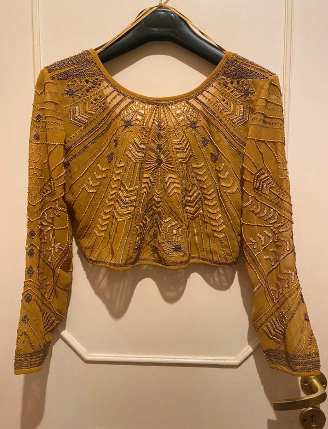 Mustard blouse with geometric embroidery in pearls and sequins, hanging on a black hanger over a beige door.