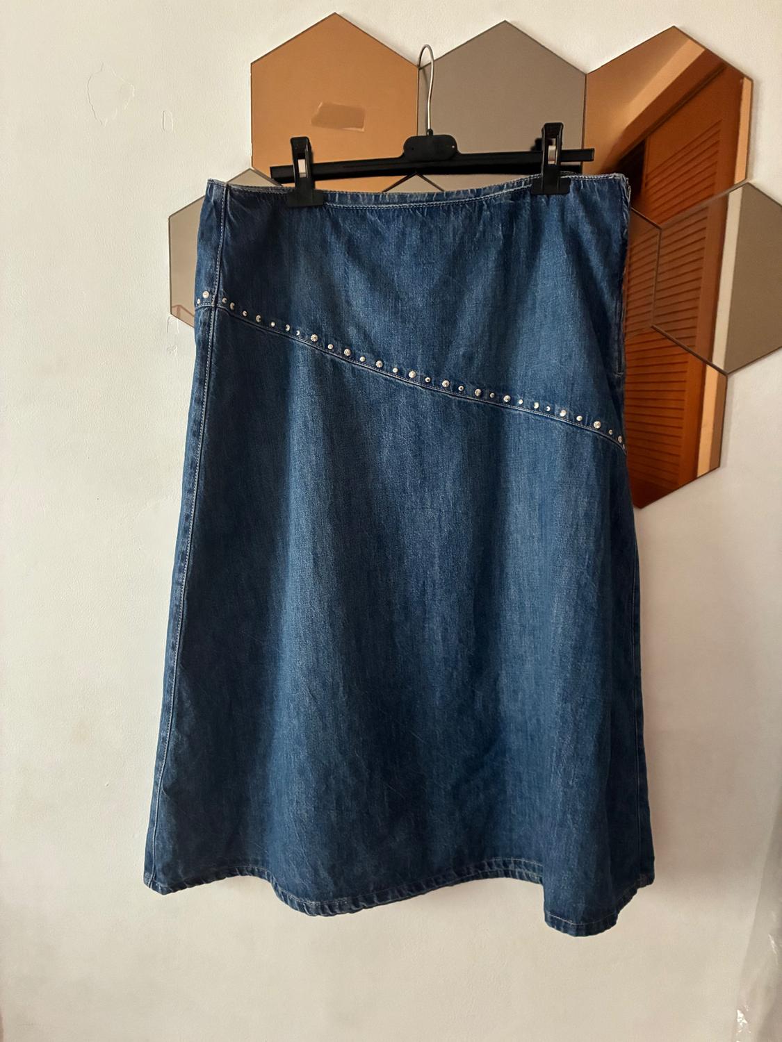 Blue denim skirt with diagonal stud detailing, hanging on a hanger in front of a hexagonal mirror on the wall.