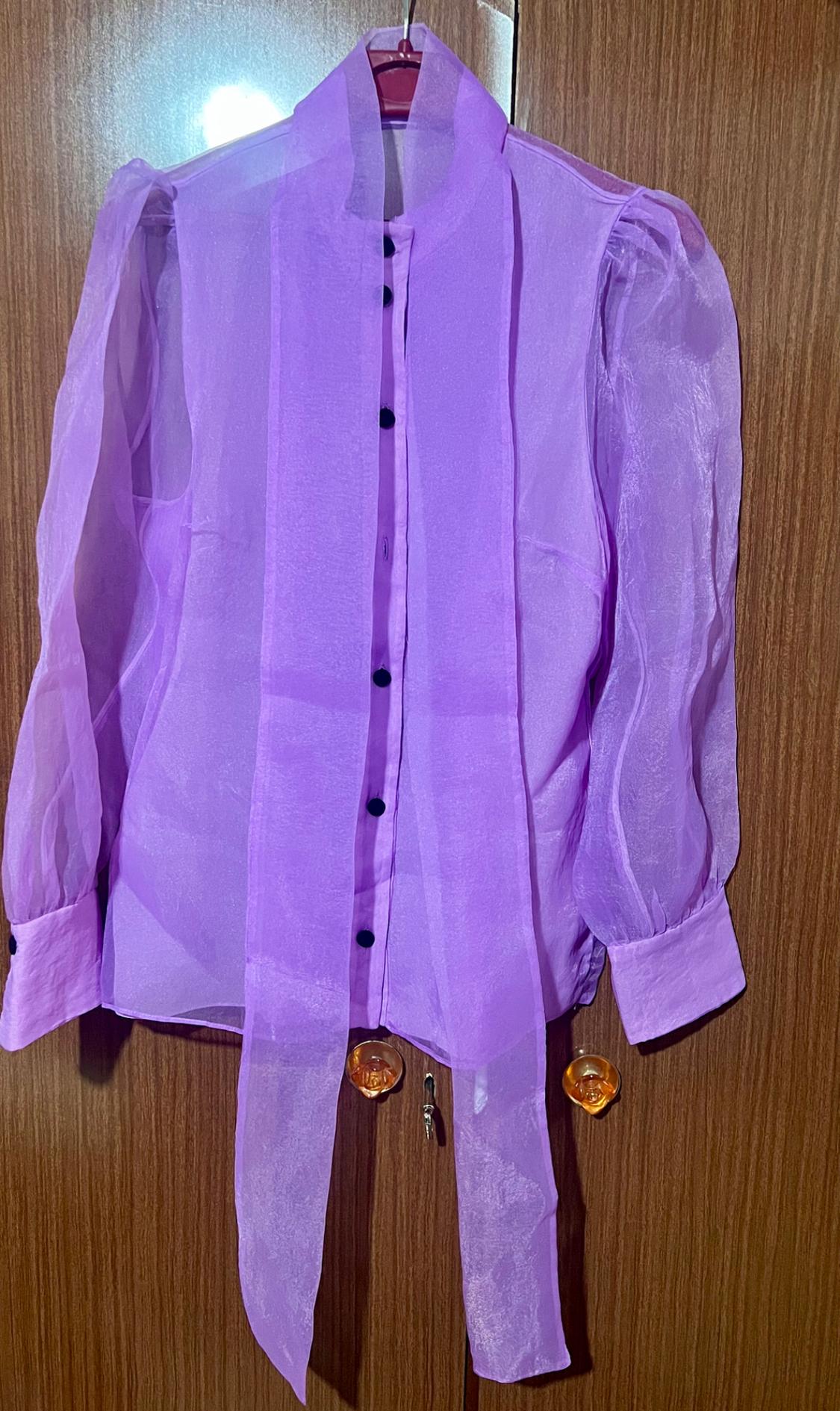 Lilac sheer blouse with long puff sleeves and black buttons, hanging on a hanger over a wooden door.