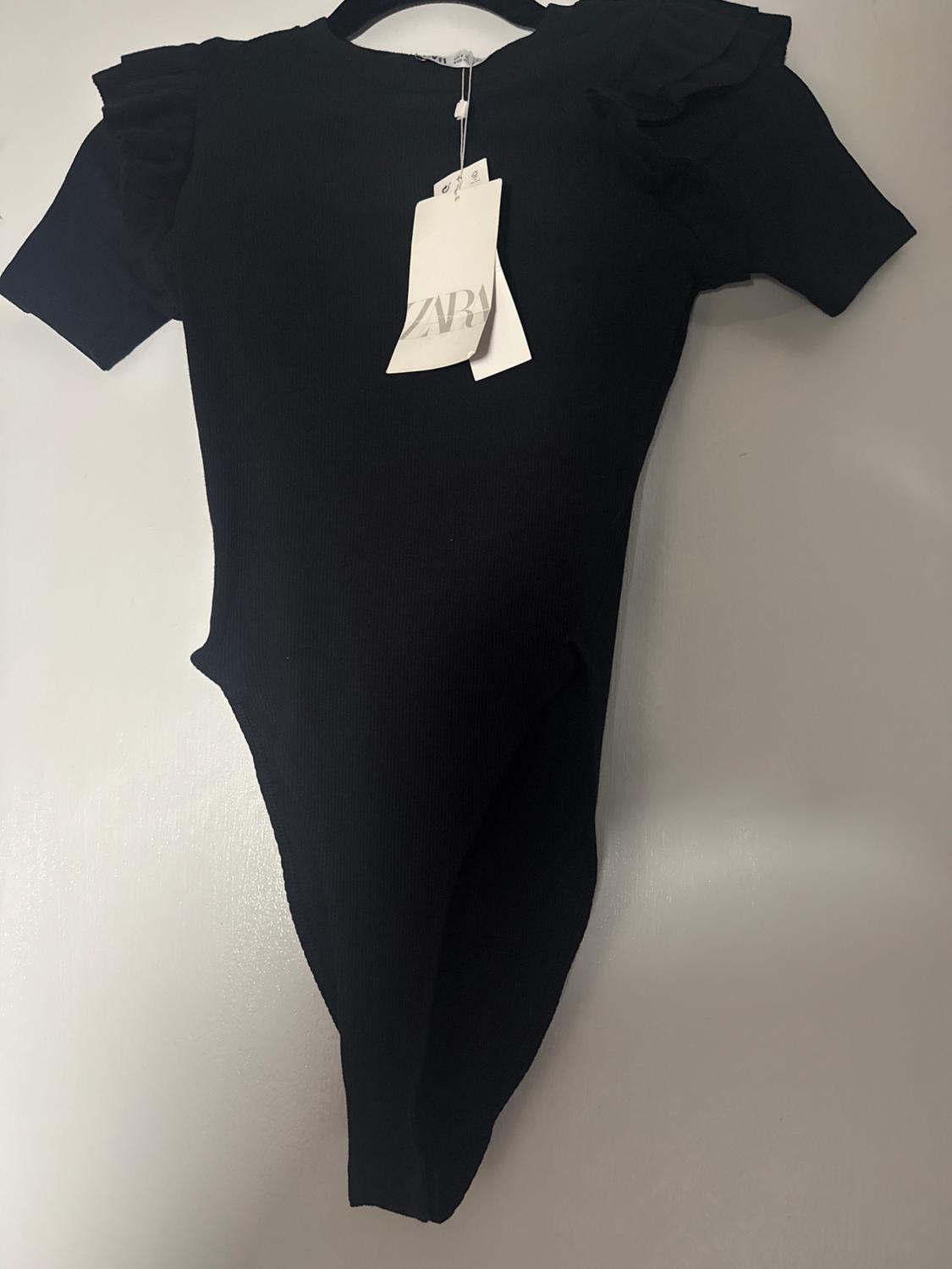 Black short-sleeved bodysuit with ruffles on the shoulders, hanging on a hanger. Visible tag with the text "ZARA".