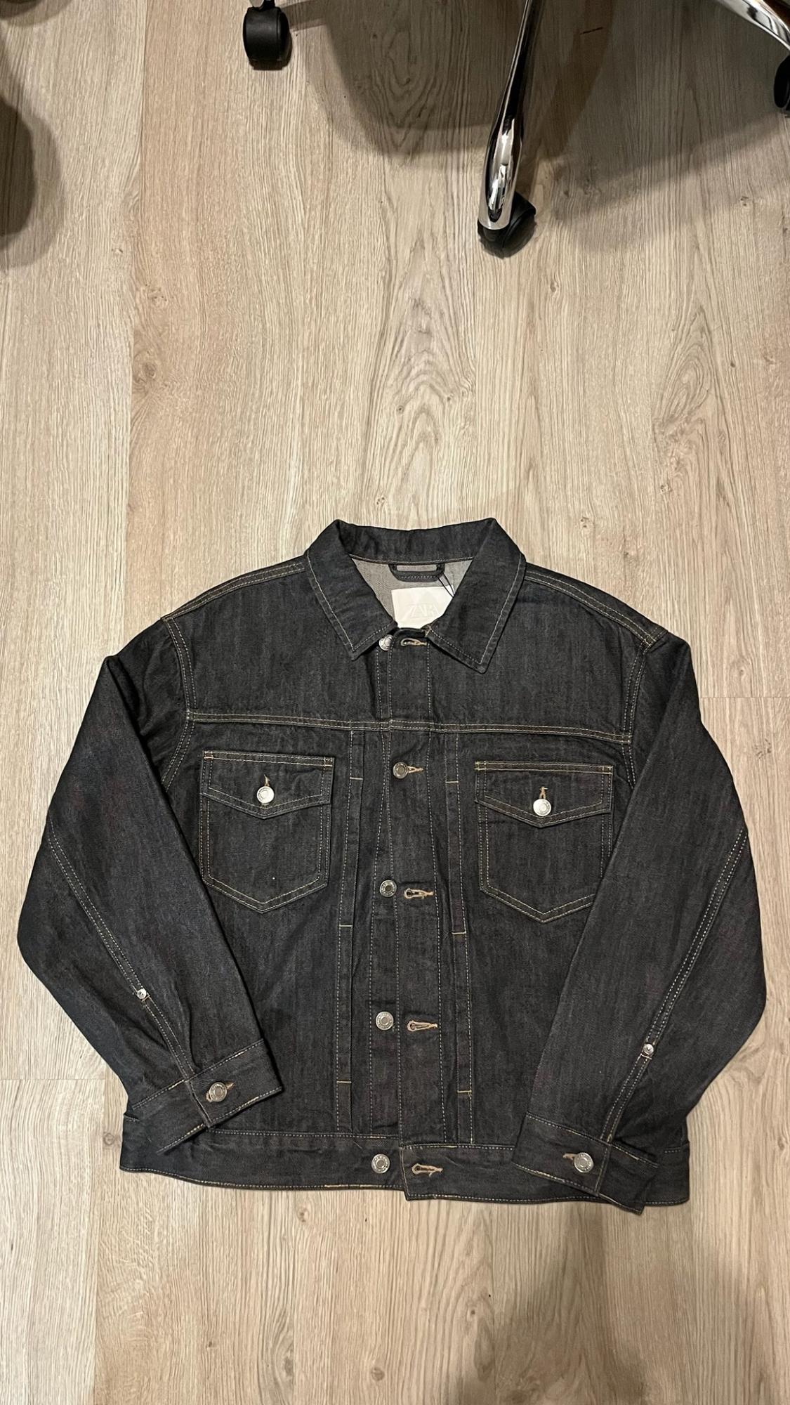 Dark denim jacket on wooden floor. It has pockets, buttons and a label with the text ZARA. Chair wheels at the top.