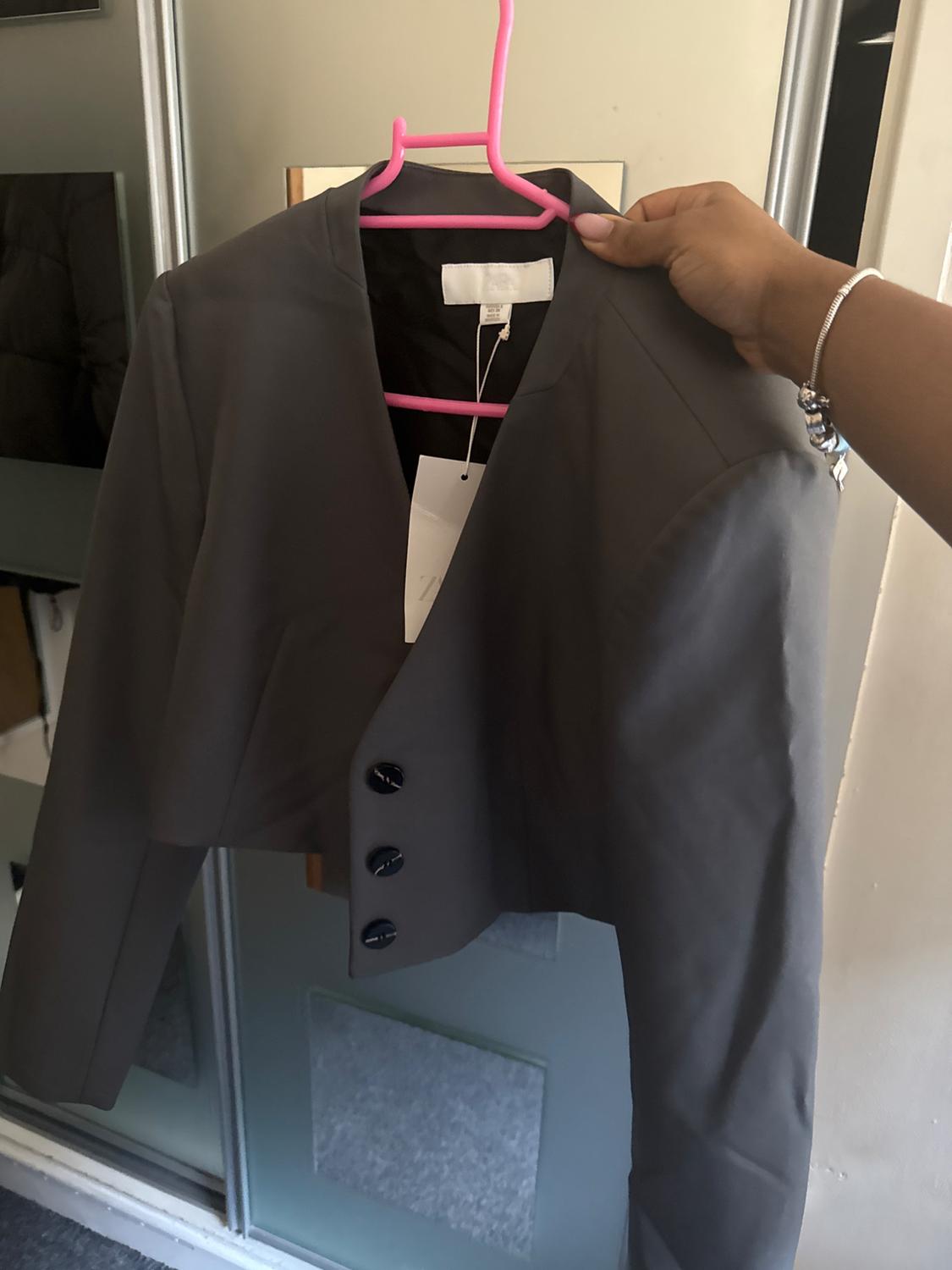 Dark grey jacket with three black buttons, hanging on a pink hanger. Mirror background and light-coloured wall. Hand holding the hanger.