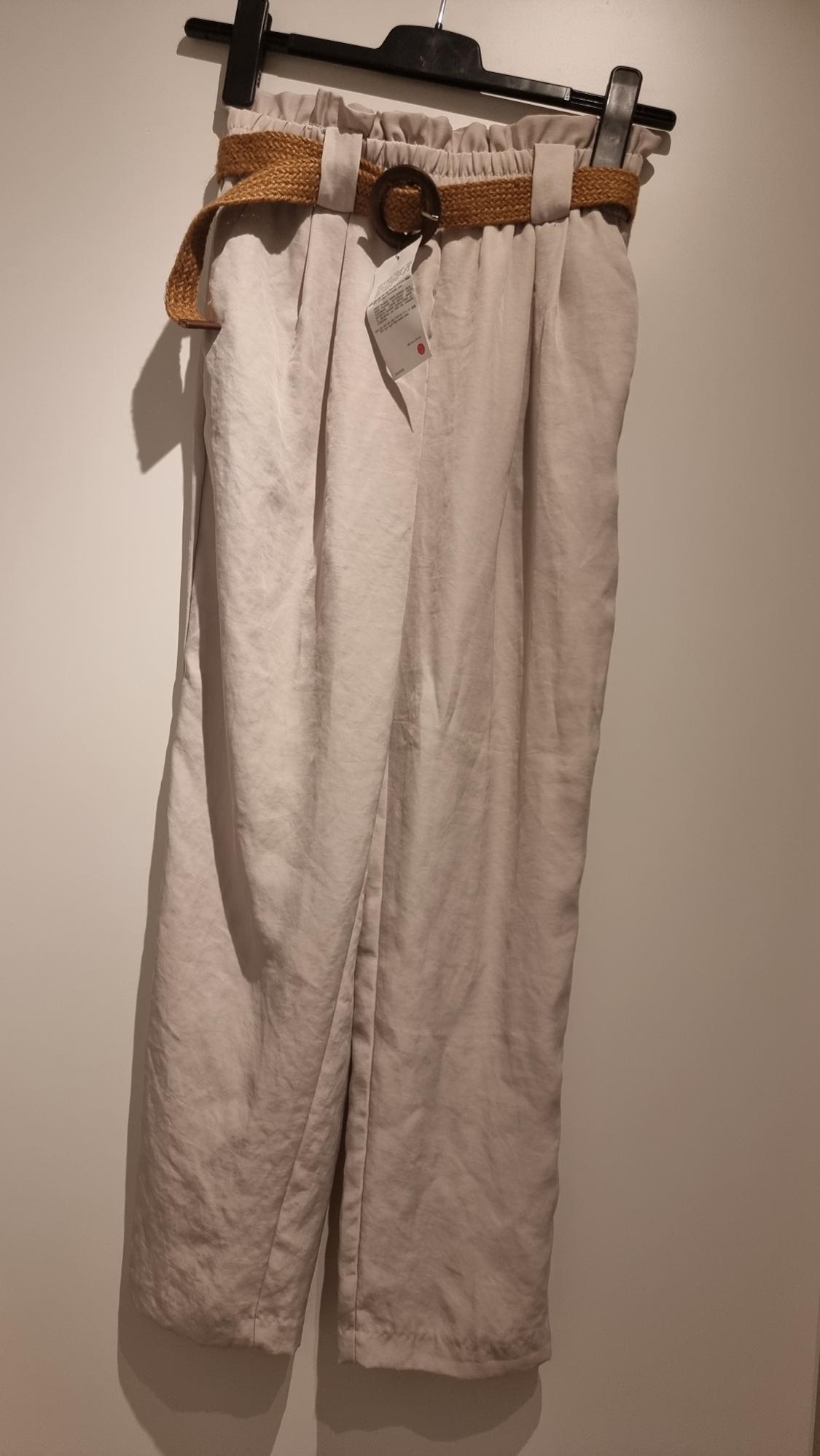 Beige trousers hanging on a black hanger, with a braided brown belt and visible tag. Illuminated white background.