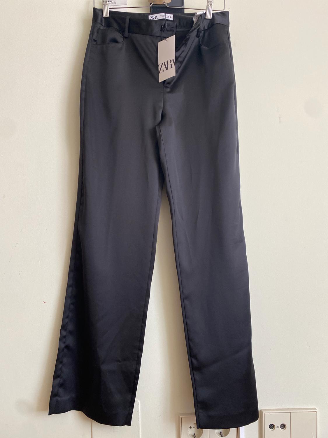 Black trousers hanging on a hanger with a visible Zara label, white background and plug sockets at the bottom of the image.