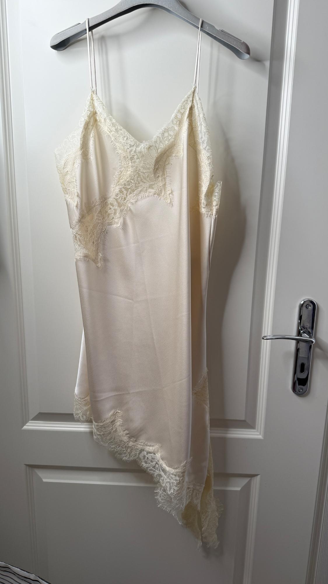 Cream-coloured satin slip dress with lace on the neckline and an asymmetric hem, hanging on a hanger on a white door.