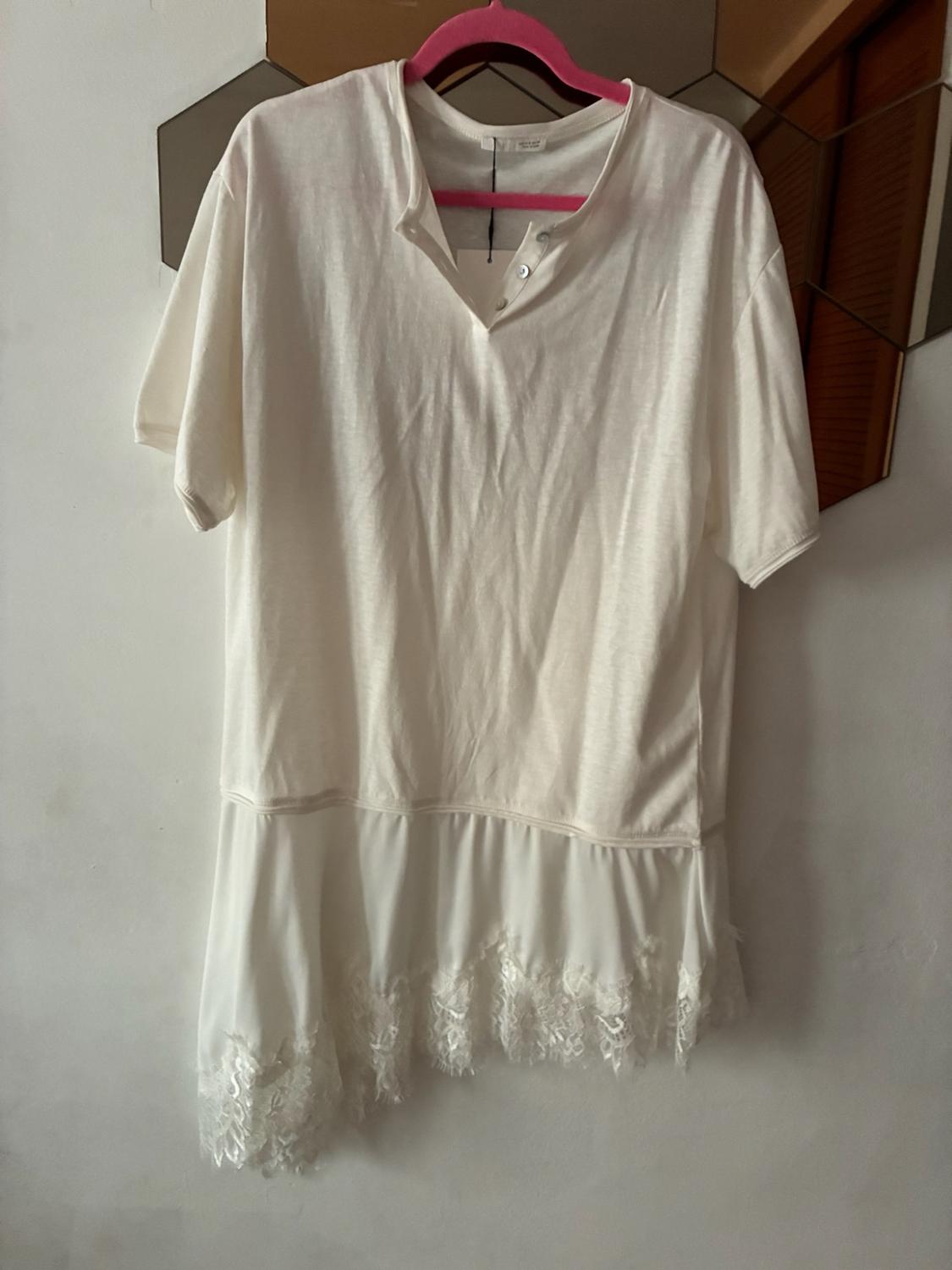 White short-sleeved T-shirt with a buttoned collar and an asymmetrical lace hem, hanging on a pink hanger against a white wall.