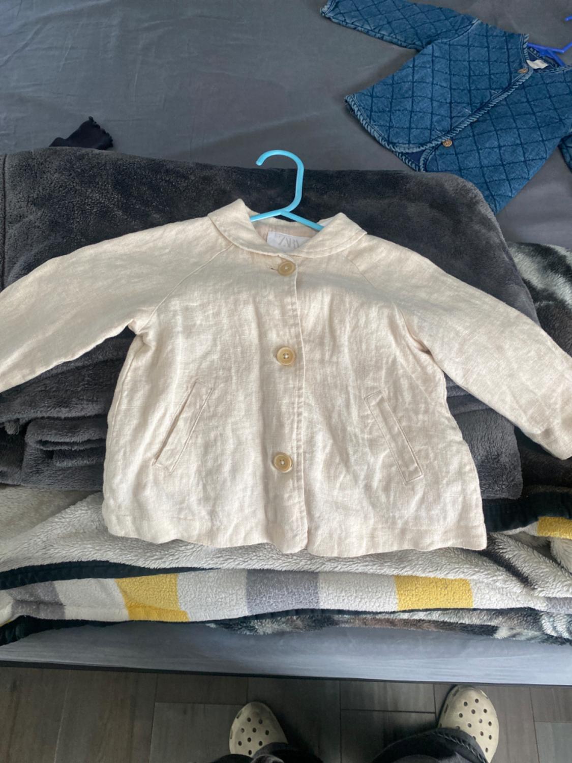 Beige linen jacket with large buttons on a grey blanket, hanging on a blue hanger. Background with another blue padded jacket.