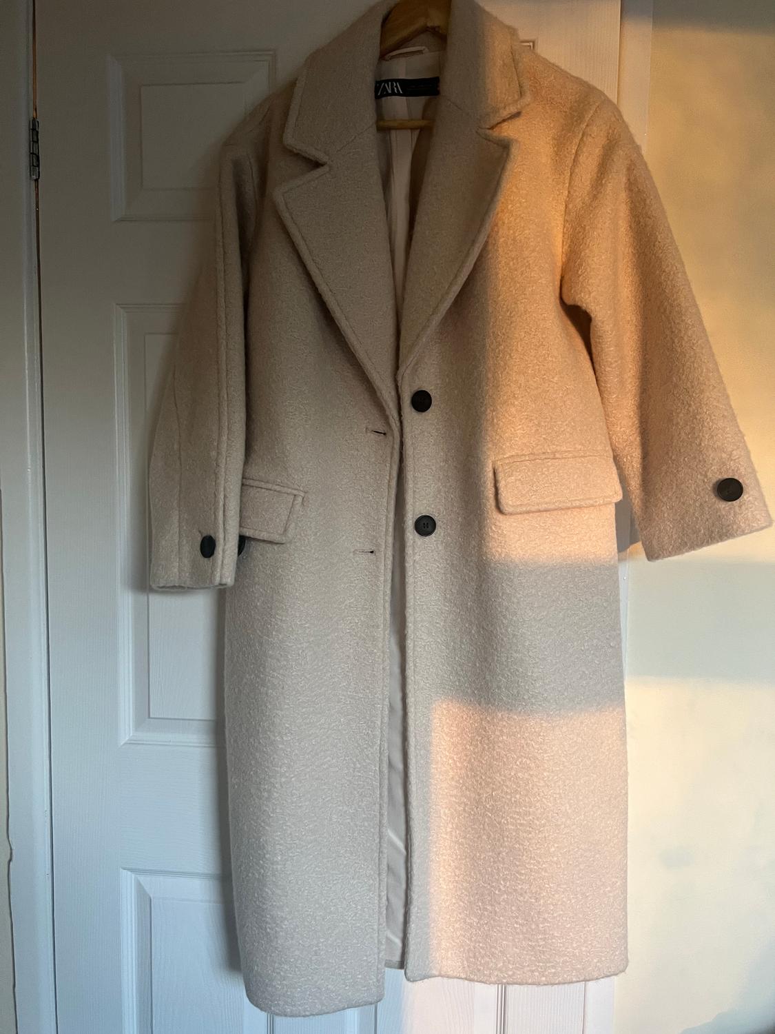 Beige coat hanging on a white door, with a wide lapel and two dark buttons. Visible label on the inside of the collar.
