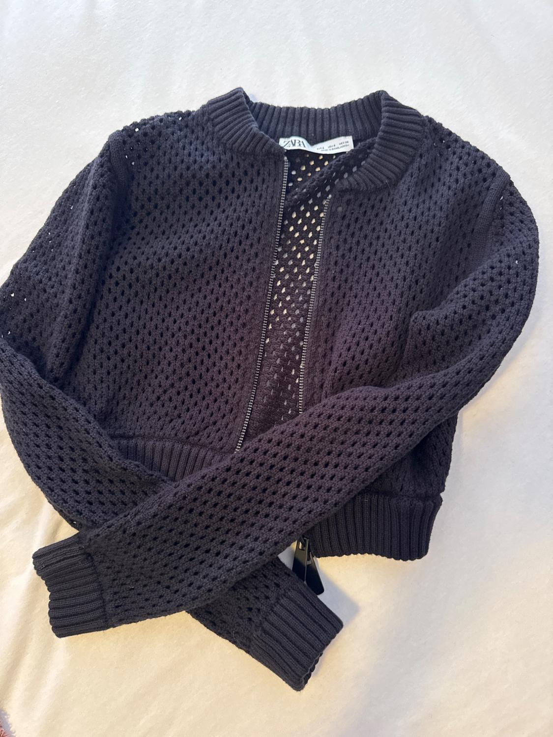 Black open-knit jumper with a zip and ribbed trims. ZARA text label. Garment with crossover sleeves on a white background.