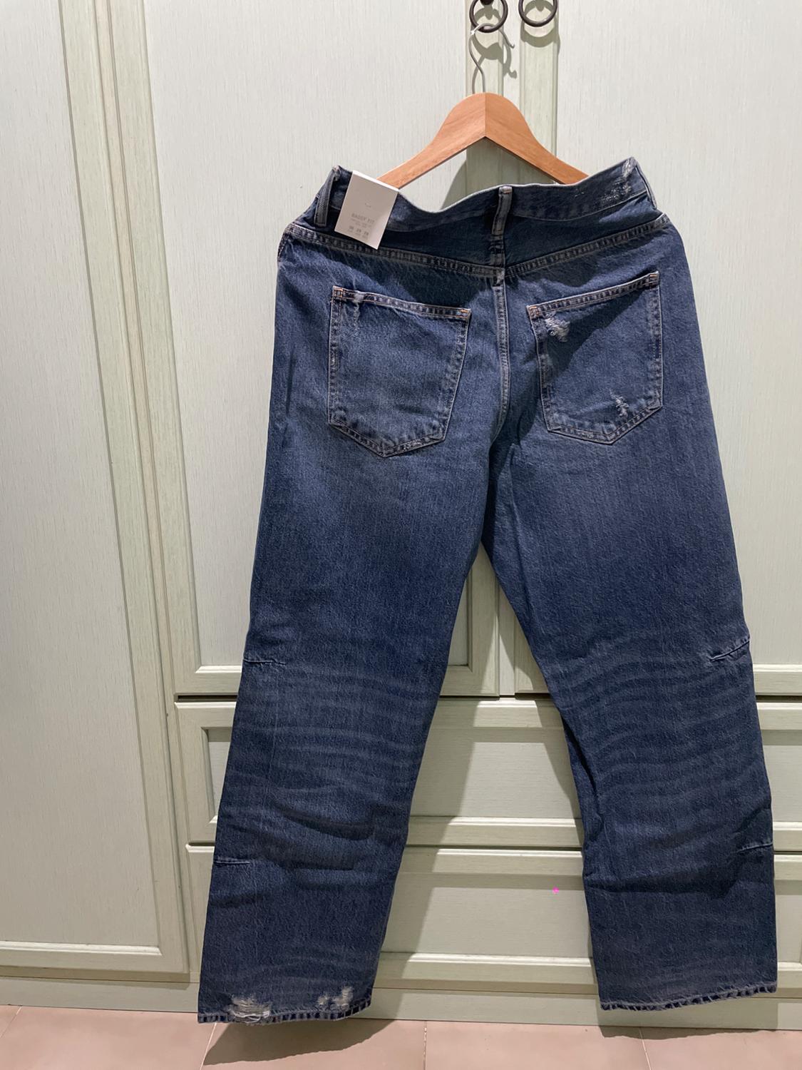 Blue jeans hanging on a clothes hanger in front of a light-coloured wardrobe. Seen from the back, showing distressed details and a BAGGY FIT label.
