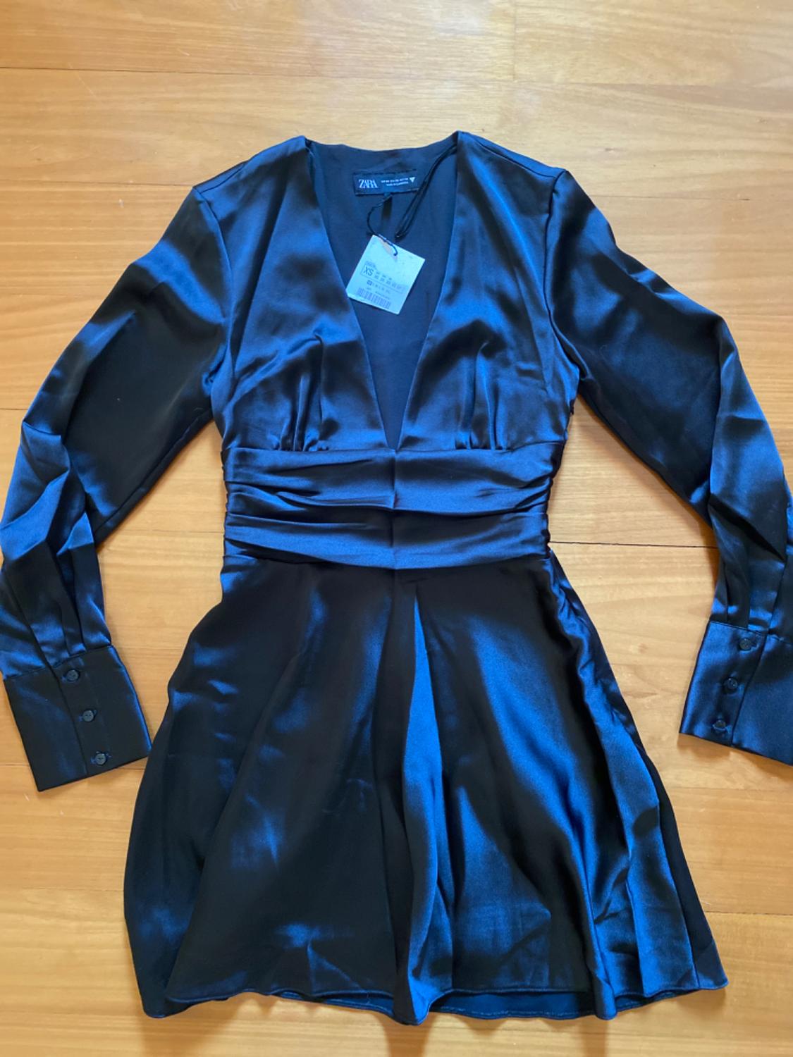 Black satin mini dress with long sleeves and V-neckline on a wooden background. Visible label on the collar.