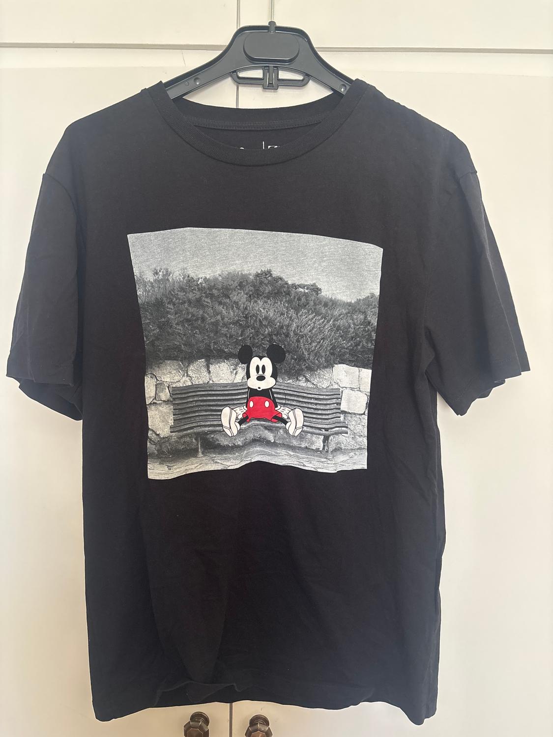 Μαύρο t-shirt με σχέδιο κινούμενου σχεδίου χαρακτήρα που κάθεται σε ένα παγκάκι. Ασπρόμαυρο φόντο με θάμνους και πέτρινο τοίχο.