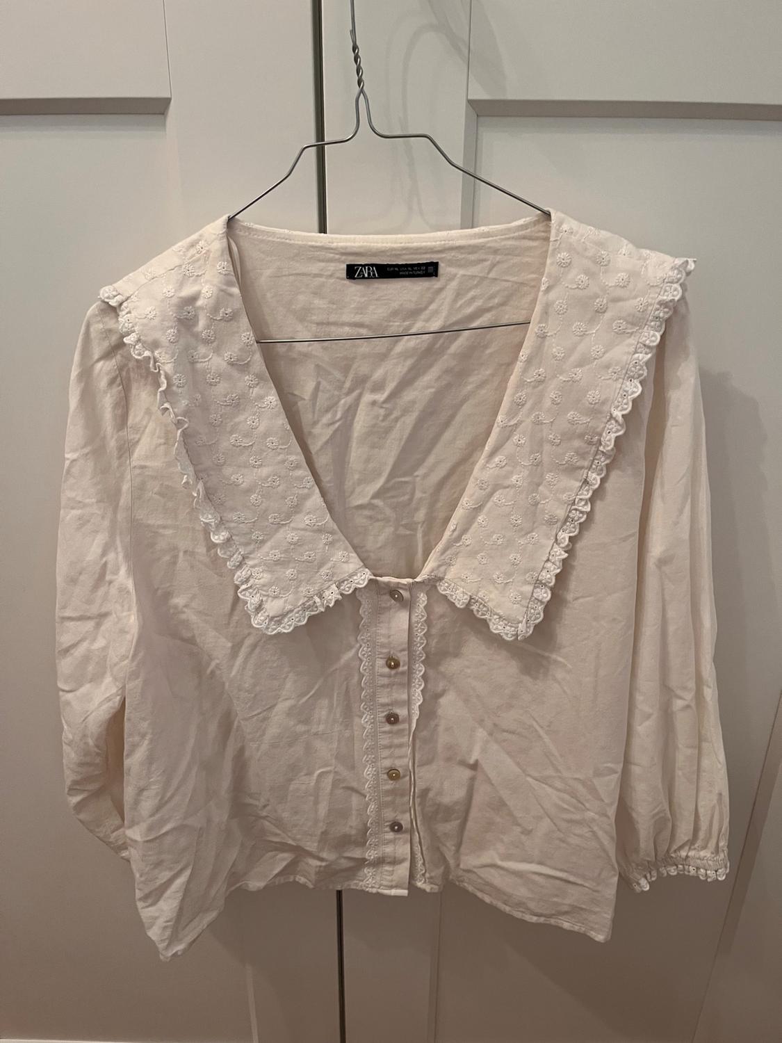 White blouse with a large embroidered collar and lace, buttoned, hanging on a coat hanger in front of a white door. Label with the text ZARA.