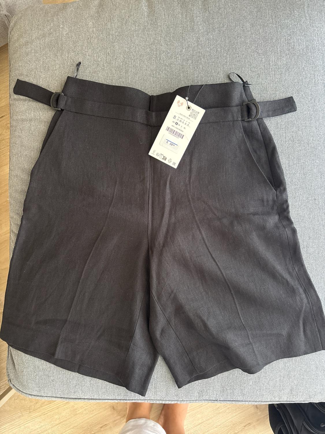 Black shorts with side buckles over grey fabric. Label with text S € 59.95. Below, bare feet on a wooden floor.
