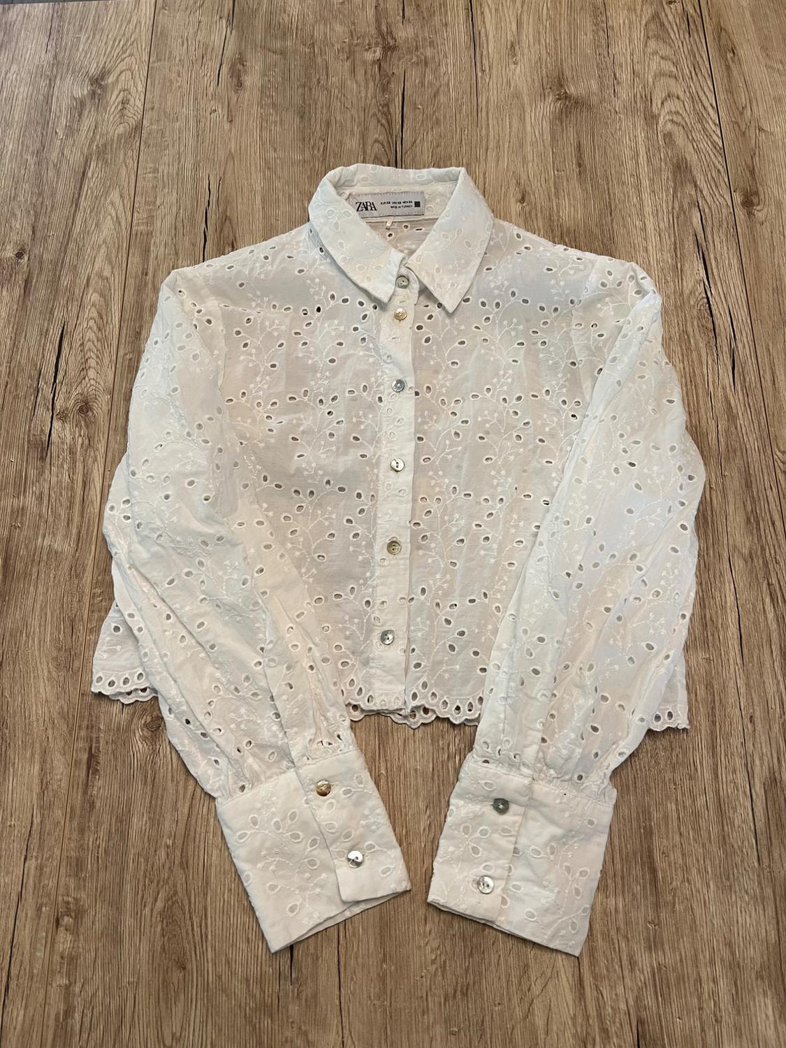 White long-sleeved shirt with openwork embroidery, buttons, and a scalloped hem. Label with ZARA text. Spread out on a wooden floor.