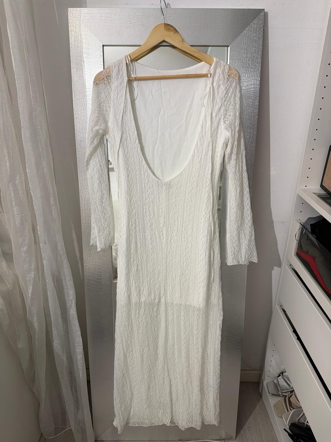 White lace dress hanging on a wooden hanger in front of a large mirror, with curtains and shelves in the background.