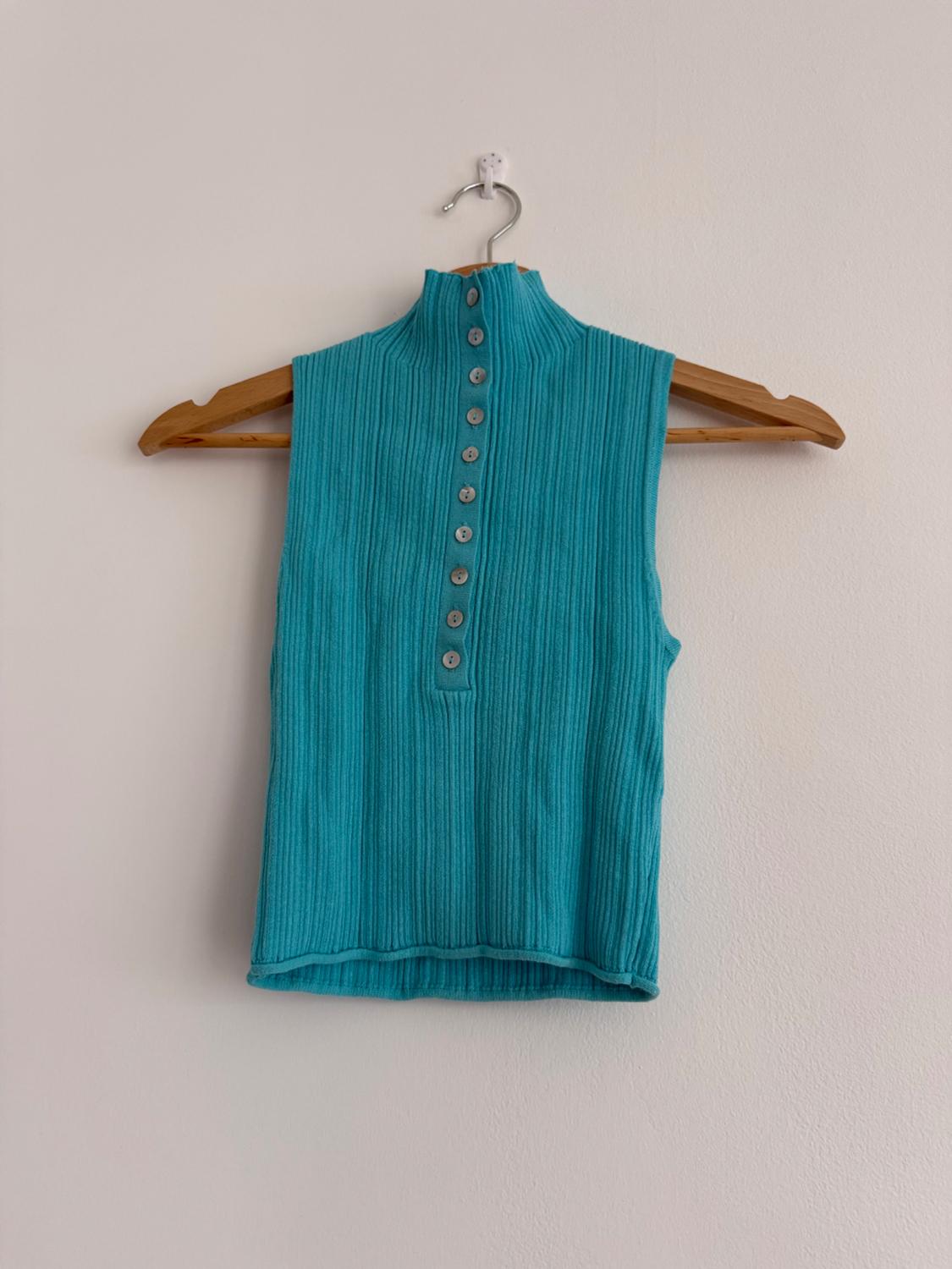 Sleeveless turquoise blue gilet with a roll neck and buttons, hanging on a wooden coat hanger against a white wall.