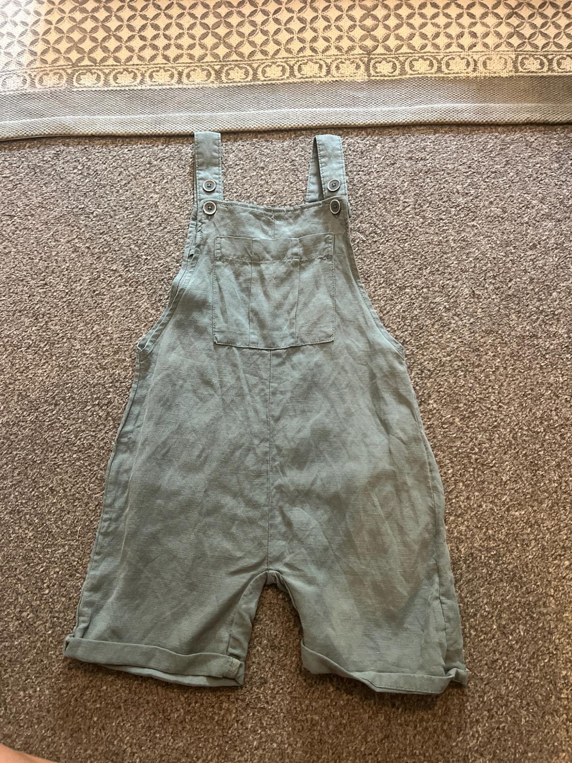 Light green dungaree shorts with buttoned straps, a front pocket and rolled hems, set on a brown rug and a geometric tapestry.