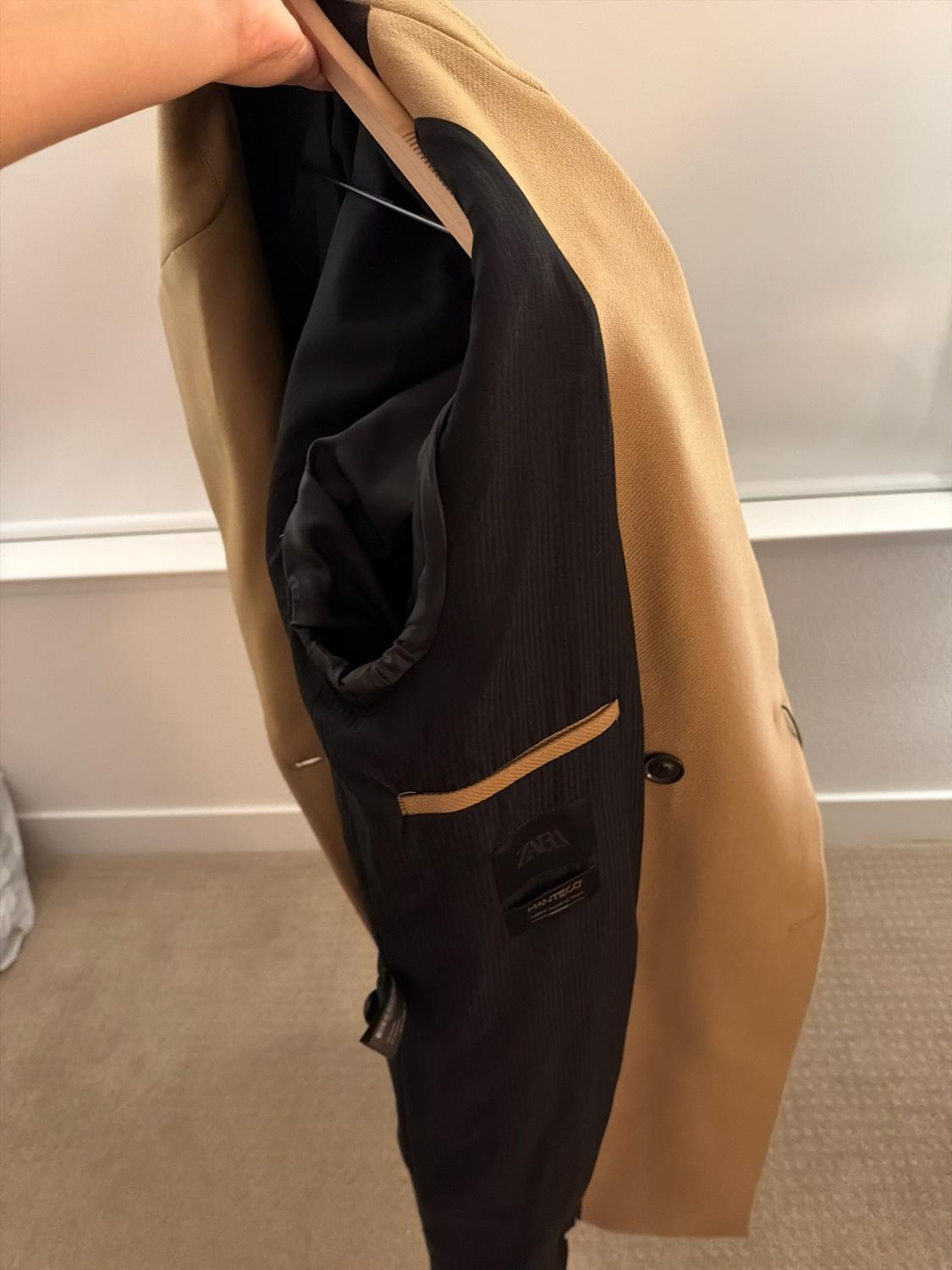 Hand holding a beige coat showing the black inner lining, a pocket, and labels with the text ZARA and MANTECO.
