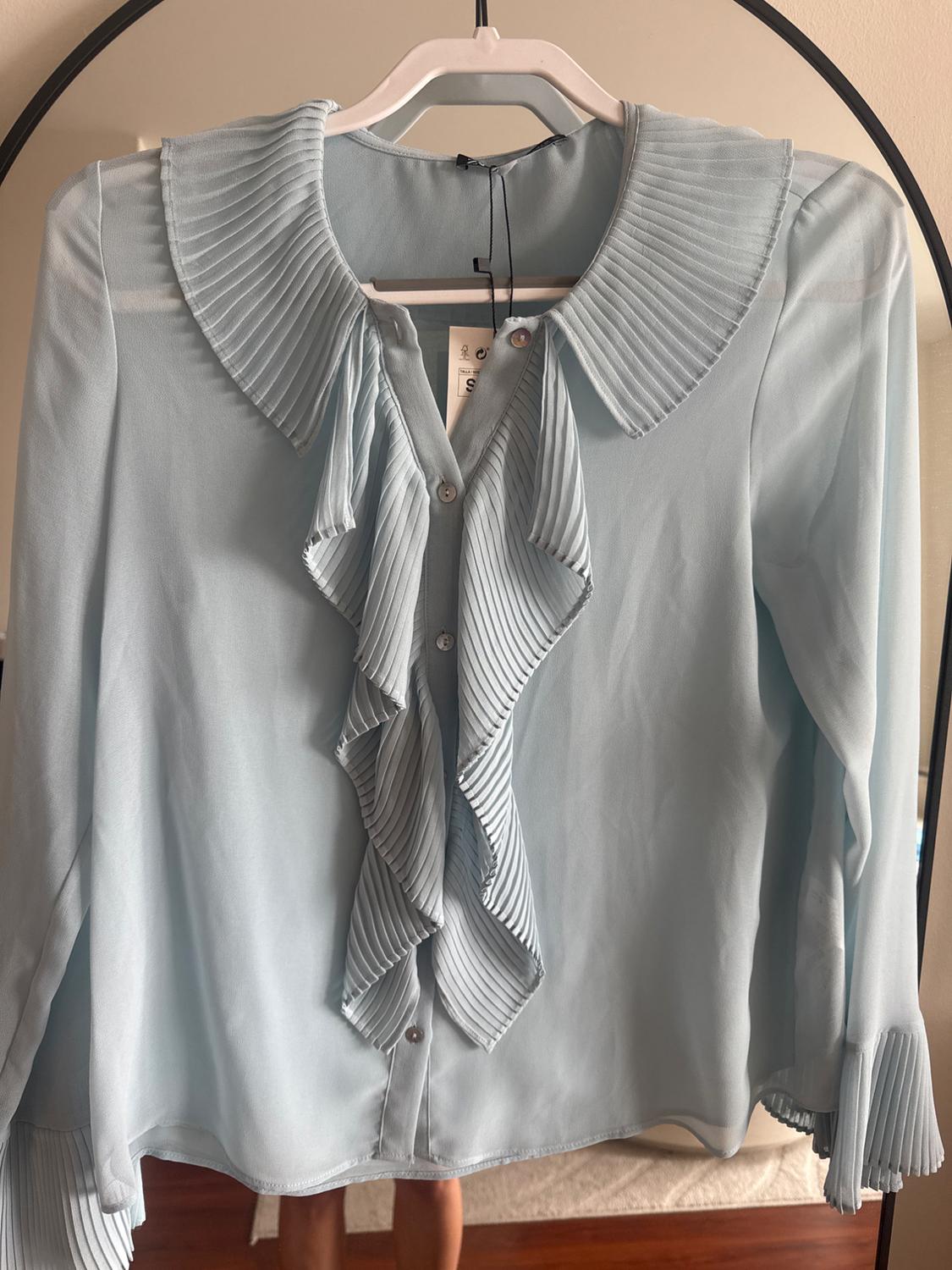 Light blue blouse with pleated ruffles on the front and collar, hanging on a white hanger. Label visible in the centre.